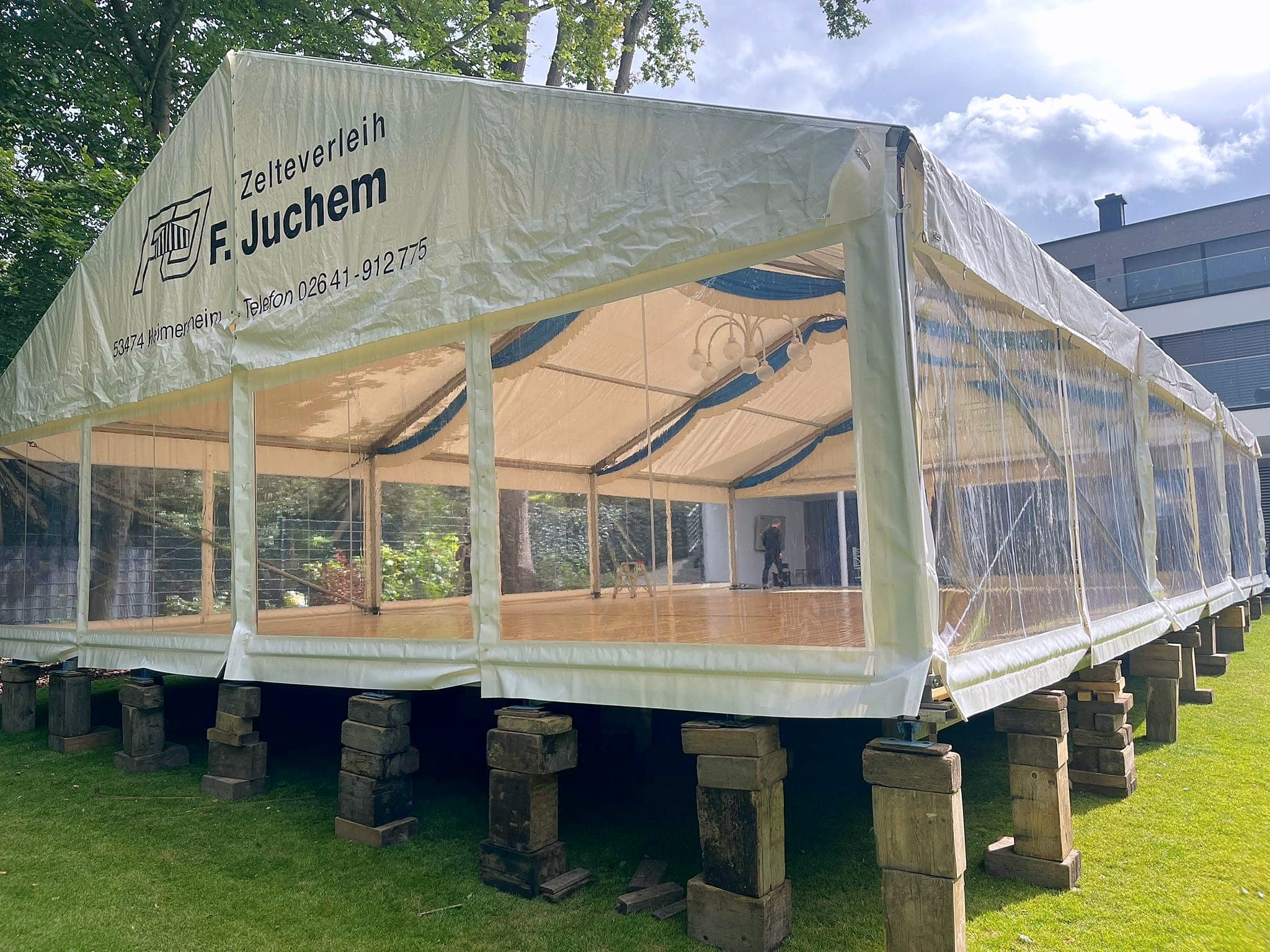 Zelt mit transparenten Seitenwänden und Juchem-Schriftzug auf einem hohen Unterbau im Garten