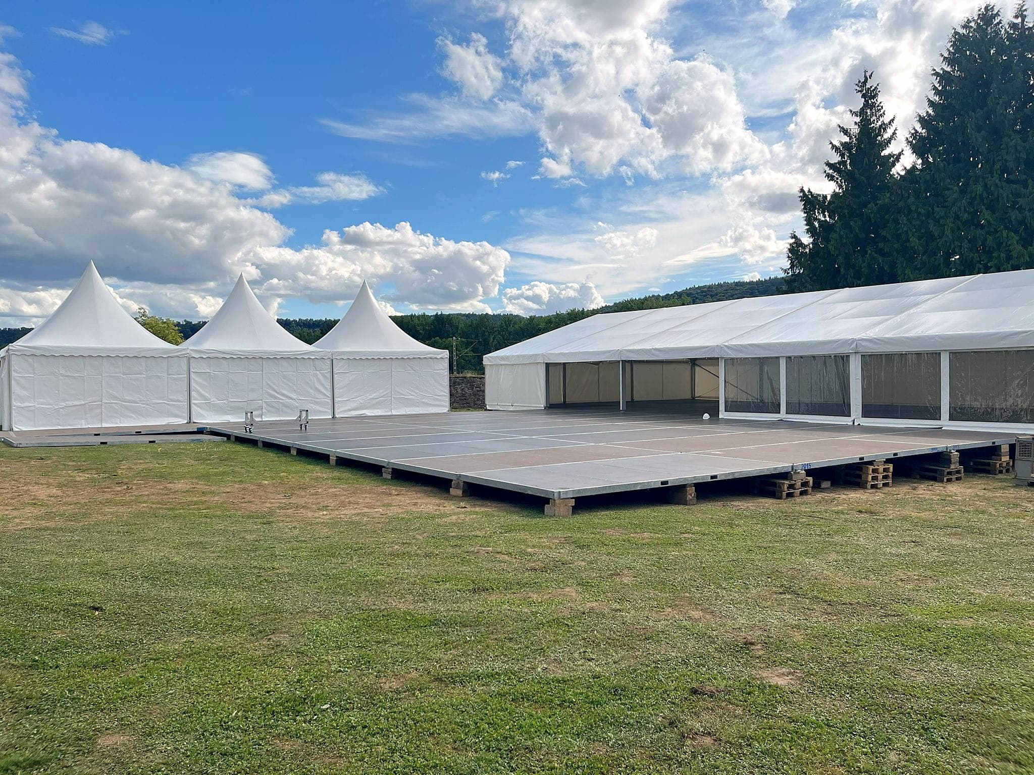 Aufbau auf einer Wiese: verlegtes Bodenpodest auf Unterbau, dahinter drei Pagodenzelte und ein Festzelt mit transparenten Seitenwänden