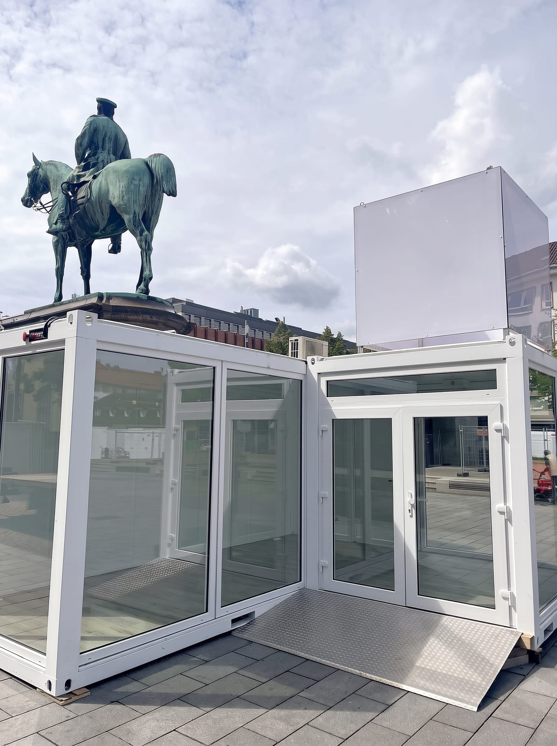 Zwei verbundene Container mit Glasfronten und Rampe auf einem Stadtplatz vor einem Reiterdenkmal