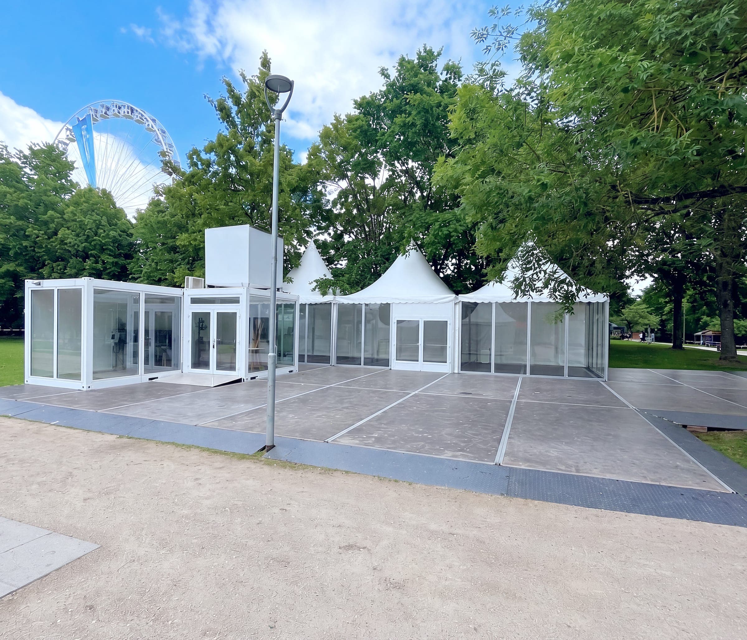 Event-Cubes mit Glasfront und ein aufgesetzter Cube neben einer Reihe Pagodenzelte mit Glaswänden auf einem Podest im Park