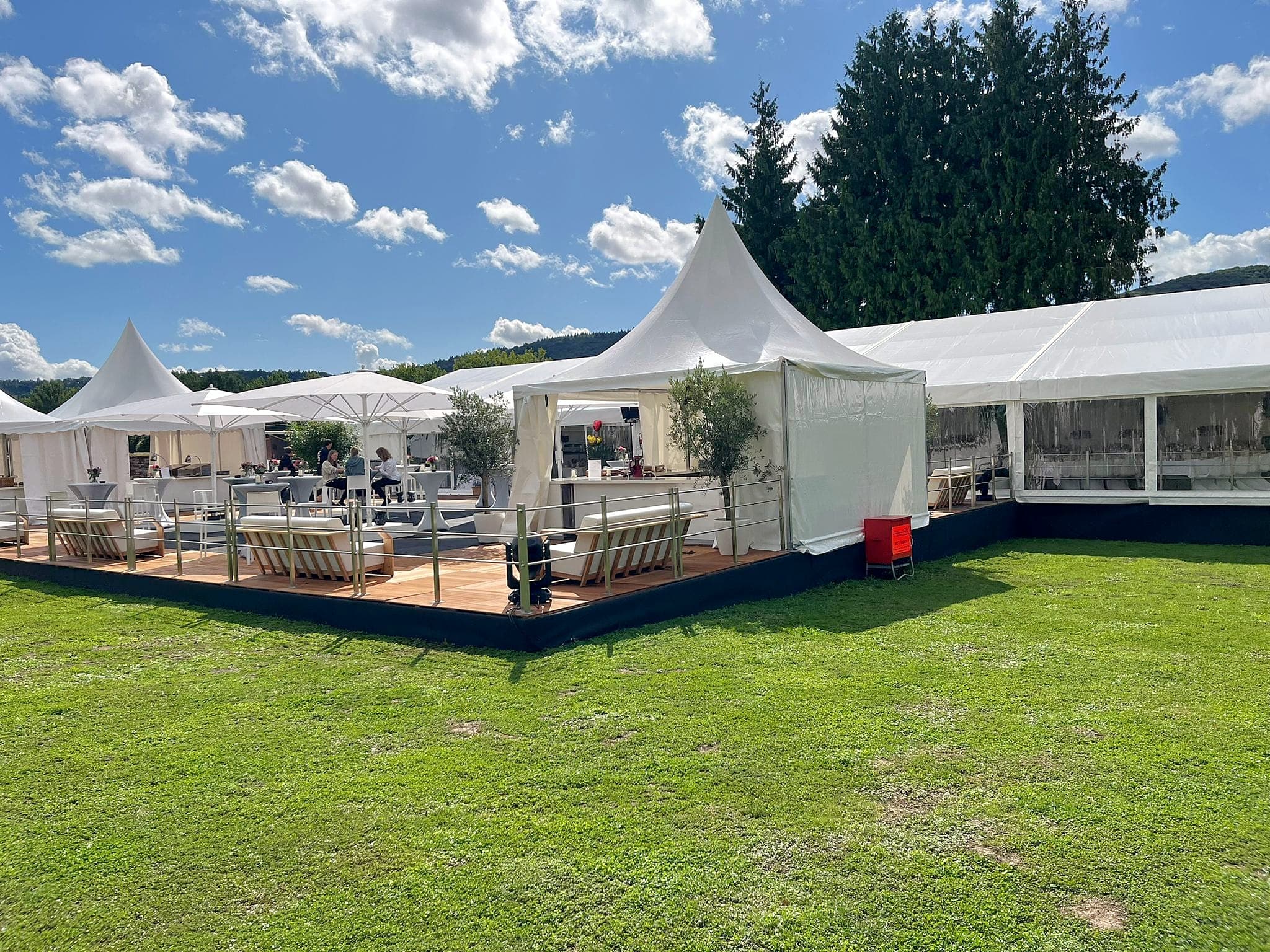 Pagodenzelte und ein Festzelt an einer Holzterrasse mit Geländer, Loungemöbeln, Stehtischen und Sonnenschirmen