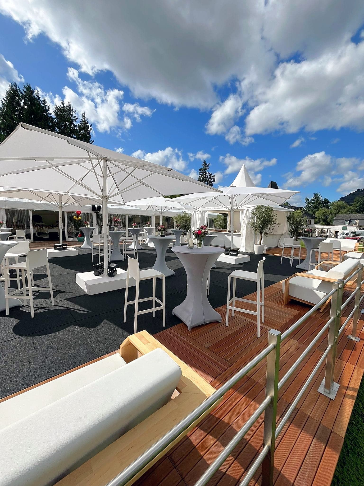 Fertig aufgebaute Terrasse mit Holzboden und Geländer, weißen Sonnenschirmen, Stehtischen und Barhockern, dahinter Pagodenzelte