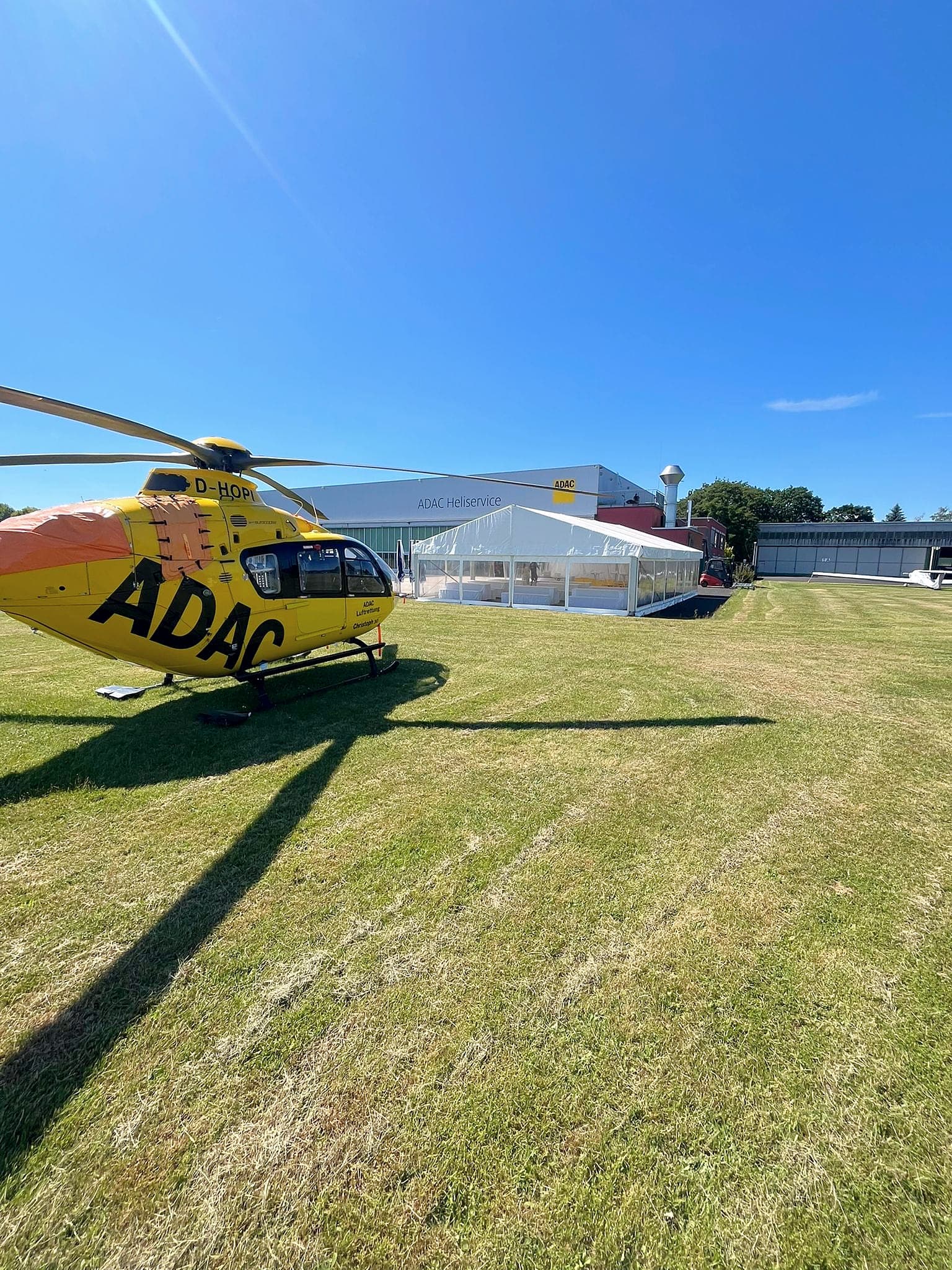 Gelber Rettungshubschrauber auf einer Wiese, dahinter ein Zelt mit transparenten Seitenwänden vor einer Hangarhalle
