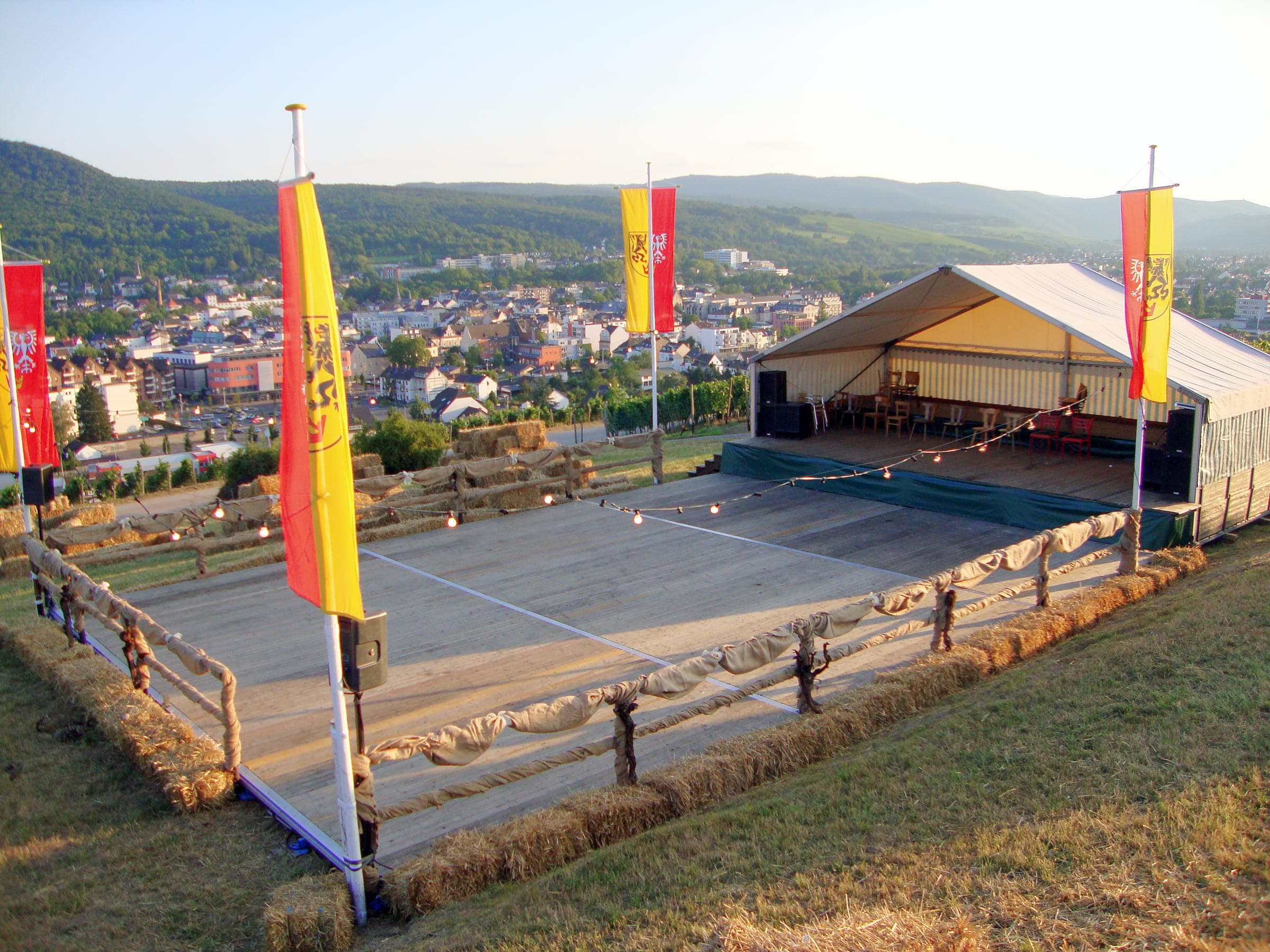 Offenes Festzelt mit großer Holzfläche am Hang, eingefasst von Strohballen und Fahnen, dahinter die Stadt im Tal