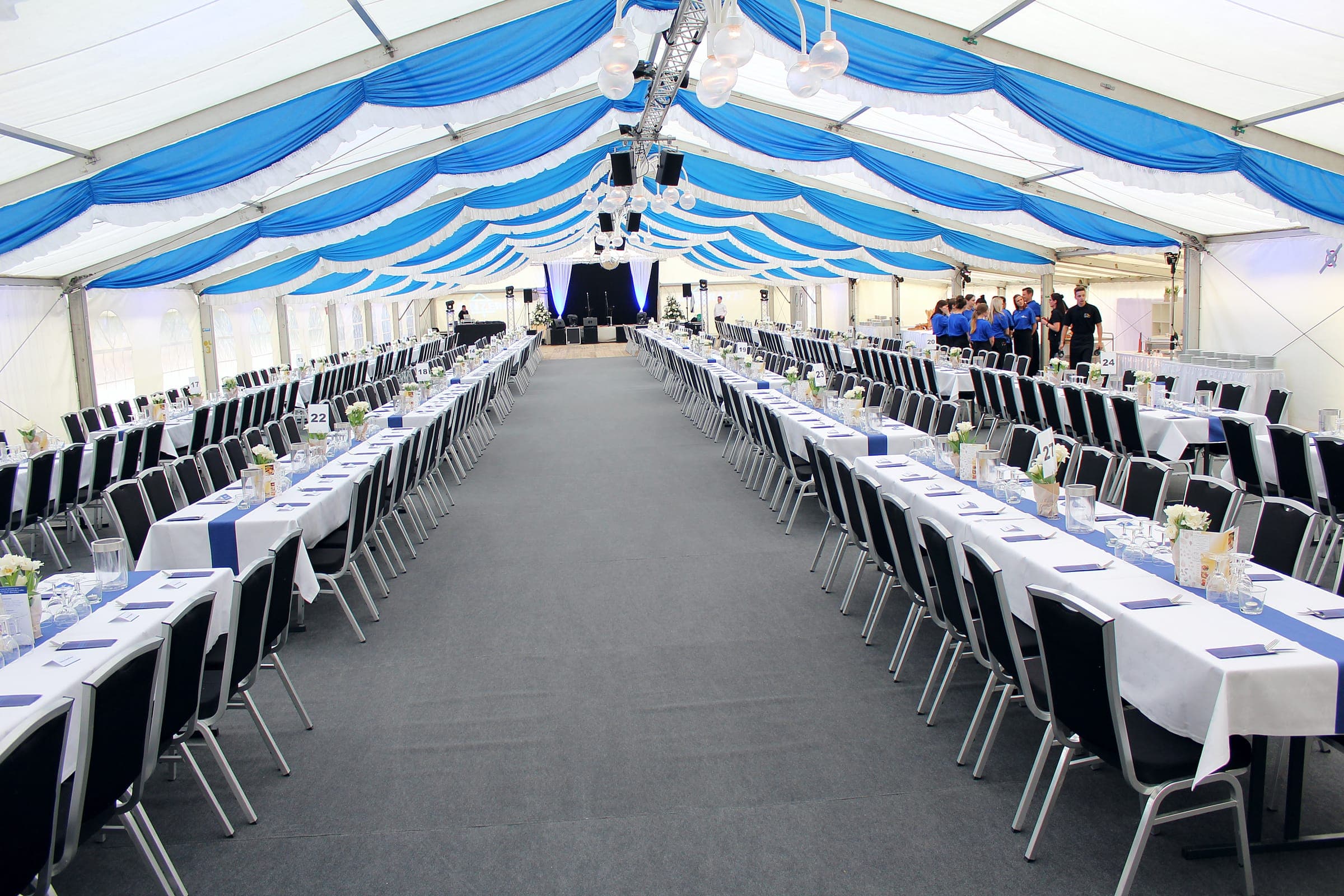 Festzelt mit blau-weißer Deckendeko, langen gedeckten Tafeln und grauem Teppichboden